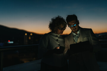 Two business people are focused and collaborating late at night, using digital devices outdoors. The warm evening cityscape provides a calm backdrop for this dedicated work session.