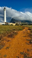 Lighthouse on the coast with mountain and sky with clouds. Yellow path on field and meadow of grass. Landscape with white beacon and trail in Sicily.