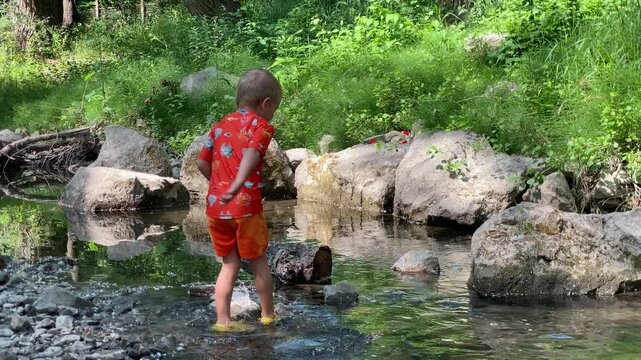 A young child, dressed in a vibrant red and orange outfit with yellow shoes, stands in a shallow creek, facing away from the viewer. The sunlight gleams on the water's surface as they navigate the roc