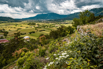 Mountain View Toward Ajdovscina From Vipava Castle: Rolling Green Hills, Wildflowers, And A Distant Town Nestled Below Steep Rocky Peaks Under A Moody Summer Sky In Western Slovenia
