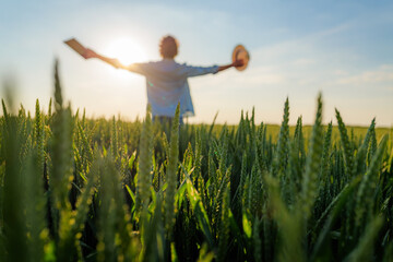 Happy farmer is holding a hat and a tablet while enjoying the sunset over a beautiful green wheat field, expressing freedom and success in agriculture