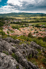 Picturesque View Of Vipava From Castle Ruins: Red-Tiled Rooftops And Church Tower Nestled In Green Valley With Blooming Flowers In Foreground And Rolling Fields In Background, Slovenia