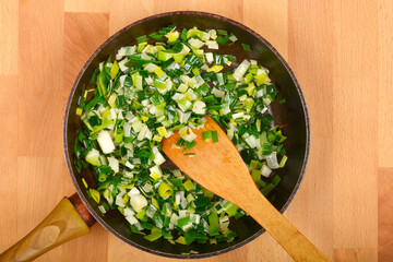 Chopped green and white leeks are being sauteed in a dark frying pan with a wooden spoon, viewed...
