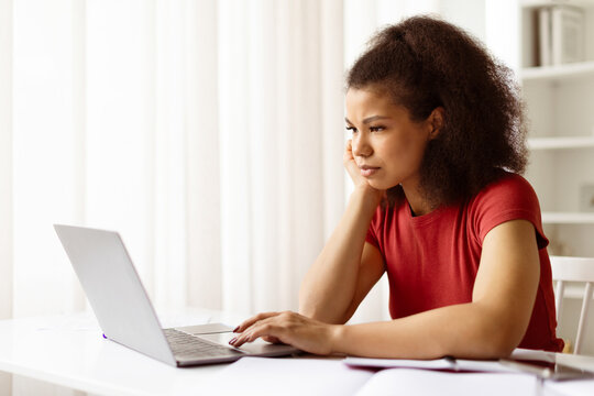 Thoughtful African American woman student looking seriously at her laptop while sitting at desk indoors, concerned or focused young black female studying with computer at home, browsing internet