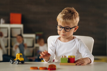 White shirt, eyewear, sitting by table and playing. Group of children are in the kindergarten