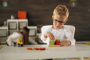White shirt, eyewear, sitting by table and playing. Group of children are in the kindergarten