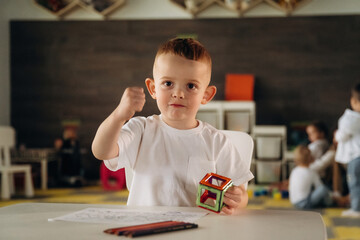 Cute boy is sitting by the table and playing. Group of children are in the kindergarten