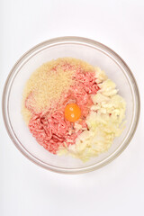 Top-down shot shows a clear glass bowl containing raw ground meat, uncooked rice, chopped onions, and a cracked egg yolk, ready for mixing to prepare stuffed peppers