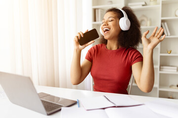 Joyful African American student woman singing into smartphone, wearing headphones and enjoying...
