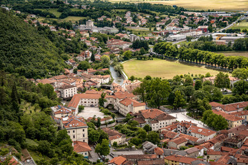 Picturesque View Of Vipava From Castle Ruins: Red-Tiled Rooftops And Church Tower Nestled In Green Valley With Blooming Flowers In Foreground And Rolling Fields In Background, Slovenia