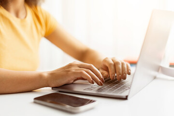 Hands of young African American woman typing on laptop in office environment, closeup of fingers rest on keyboard, while smartphone lies nearby on the white desk, ideal for studying or remote work