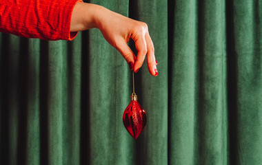 Female hand in red sleeve holds a shiny red Christmas ornament against a green curtain. Minimal and...