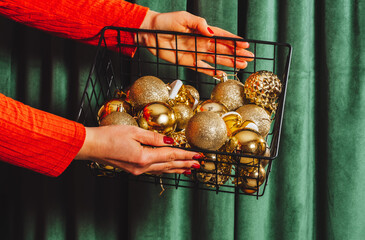 Hands holding a black basket filled with shiny gold Christmas ornaments against green velvet curtains. Festive and elegant decor concept.