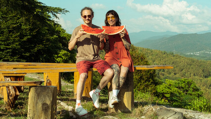 Fototapeta premium Smiling couple enjoys juicy watermelon slices while sitting on a bench in the mountains. A cheerful summer moment filled with nature, sunshine, and fun.