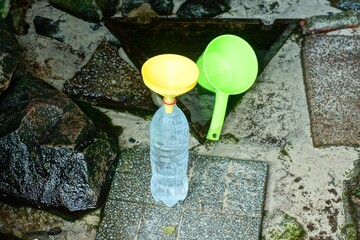 a plastic transparent white bottle with clean cold water and a yellow watering can stands on a stone near a green round empty bath ladle with a handle near a spring from under the ground