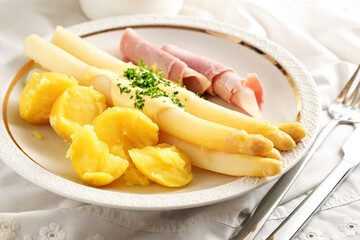 White asparagus, potatoes and boiled ham with hollandaise sauce and chive garnish on a festive white plate with napkin and cutlery, seasonal dish, selected focus