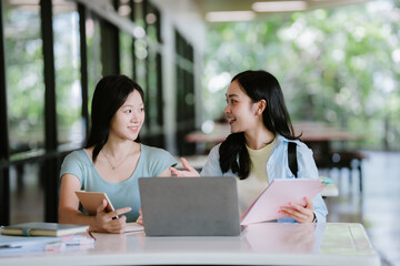 Two cheerful Asian female students working on a school project using a shared laptop and tablet in a library.
