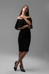 Elegant woman poses thoughtfully in an off-shoulder black dress against a neutral backdrop