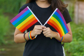 A woman wearing a black T-shirt holds two rainbow flags crossed at chest level. She wears a black wristband and a rainbow wristband, which is a symbol of lgbt pride.