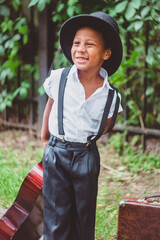 a boy dressed in the style of the 20s laughs and holds a guitar by the neck behind his back, looking to the side