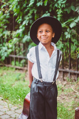 a happy boy dressed in the style of the 20s holds a guitar behind his back by the neck looking to the side