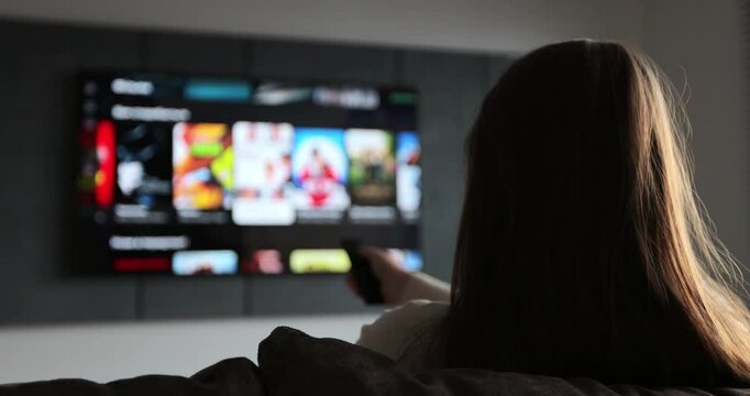 Woman selects tv channels with remote control at living room on sofa. Woman controls TV using a modern remote control.