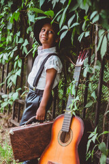 boy dressed in the style of the 20s stands next to a guitar holding a suitcase in his hand looking up