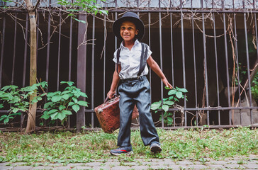 A boy dressed in the style of the 20s with a suitcase in his hand against the background of a fence