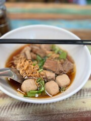 Authentic Thai beef noodle soup, a rich and aromatic broth with tender beef slices, beef balls, and various herbs, topped with crispy fried garlic and fresh greens, served with chopsticks.