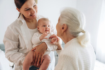 Standing, looking at each other. Mother, grandmother and little baby child on hands indoors