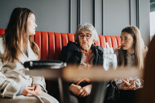 A senior woman sharing a moment with two young women, sitting in a cafe booth. The group engages in meaningful conversation, evoking themes of family, connection, and generational dialogue.