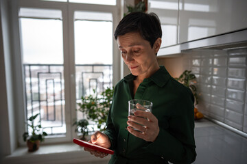 Nervous worried mature woman checking phone at home, overwhelmed by content, drinking water. Online negativity, life updates strain, personal worries, modern connectivity burden, solitude with tech