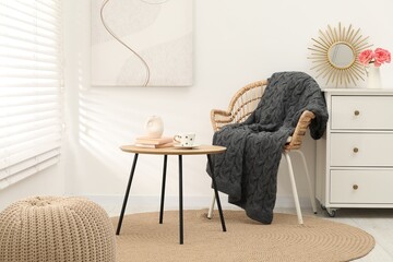 Wicker chair with gray woolen blanket, books, vase and cup on wooden table indoors