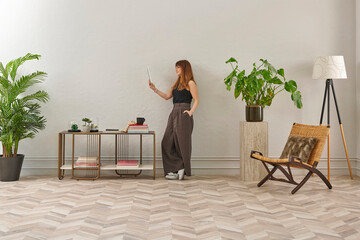 A woman with glasses sits in a woven chair, looking at a phone. She wears a black top, dark pants, and cozy grey slippers.
