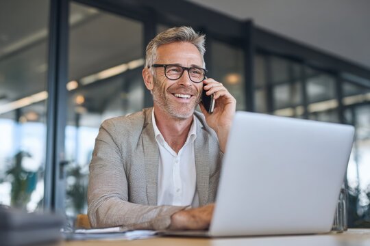 A smiling businessman using a mobile phone while sitting at a table with a laptop in a modern office.