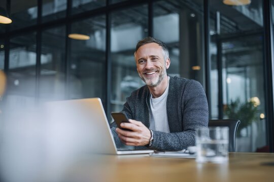 A smiling businessman using a mobile phone while sitting at a table with a laptop in a modern office.