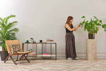 A woman in a black top and dark wide leg pants waters a large plant. The plant sits in a dark pot on a light-colored pedestal.