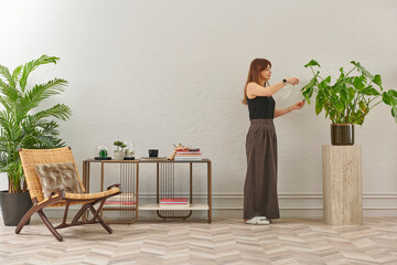 A woman in a black top and dark wide leg pants waters a large plant. The plant sits in a dark pot on a light-colored pedestal.
