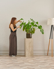 A woman in a black top and dark wide leg pants waters a large plant. The plant sits in a dark pot on a light-colored pedestal.