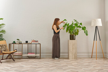 A woman in a black top and dark wide leg pants waters a large plant. The plant sits in a dark pot on a light-colored pedestal.