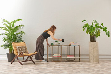 A woman in a black top and dark wide leg pants waters a large plant. The plant sits in a dark pot on a light-colored pedestal.
