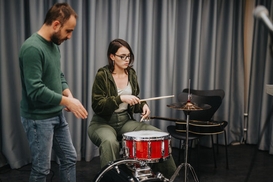 A music instructor teaching a student how to play drums during a band session in a studio. The student is seated, holding drumsticks, while the instructor demonstrates and provides guidance.