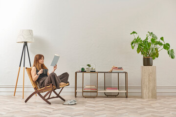 A woman is reading a book at home, looking at the phone and tablet. Decorative interior style and vase of plant.
