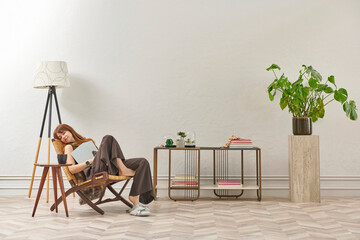 A woman is reading a book at home, looking at the phone and tablet. Decorative interior style and vase of plant.