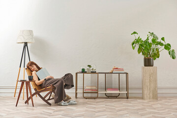 A woman is reading a book at home, looking at the phone and tablet. Decorative interior style and vase of plant.