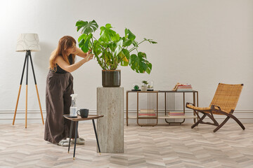 A woman in a black top and dark wide leg pants waters a large plant. The plant sits in a dark pot on a light-colored pedestal.