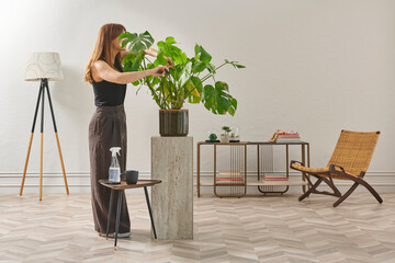 A woman in a black top and dark wide leg pants waters a large plant. The plant sits in a dark pot on a light-colored pedestal.