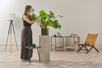 A woman in a black top and dark wide leg pants waters a large plant. The plant sits in a dark pot on a light-colored pedestal.