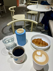 Stylish café breakfast with cappuccino, black coffee, croissant and water on white table