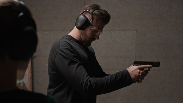 Over the shoulder view of woman in earmuffs listening to cheerful young male instructor wearing goggles showing gun stance at shooting range